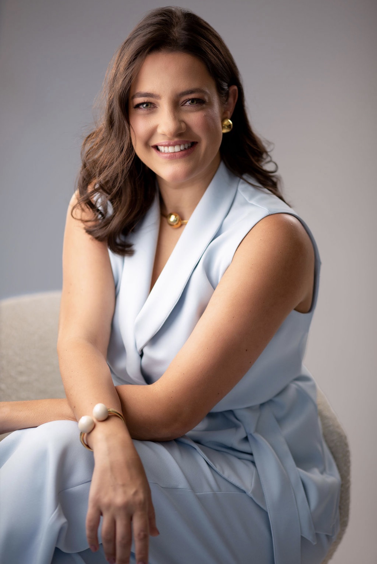 Dra. Nayara Curti, médica neurologista, sentada e sorrindo, em traje profissional azul claro